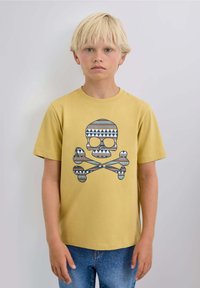 Lys blond dreng iført en sennepsgul T-shirt med et mønster af kranium og korslagte knogler, der står foran en ensfarvet lys baggrund.