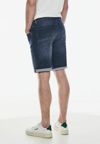 Džínové šortky v tmavě modré barvě s ohrnutými manžetami, obsahující dvě zadní kapsy. Kombinováno s bílými teniskami, které mají zelené akcenty.