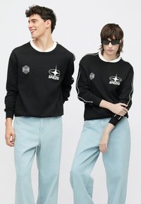 Fekete pulóverek fehér részletekkel és logókkal, világoskék, laza szabású farmerrel párosítva. Texturált szövet és kényelmes kialakítás.