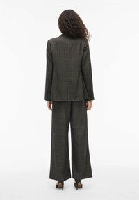 Rudas langų kostiumas su plačiomis kelnėmis ir atitinkamu švarku. Audinys turi subtilią tekstūrą. Puošia juodi mygtukai ir įpjova apykaklė.