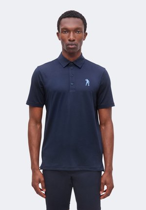 Homme portant un polo bleu marine à manches courtes avec un petit logo golfeur bleu clair sur la poitrine gauche, debout devant un fond uni.