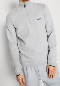 Pull léger gris clair à demi-zip avec des accents côtelés, featuring un logo "BOSS" noir sur la poitrine et des finitions noires contrastantes sur les poignets.