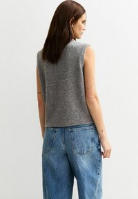 Grijze mouwloze gebreide top met een zachte textuur, gecombineerd met hoge versleten blauwe spijkerbroeken met grote achterzakken.