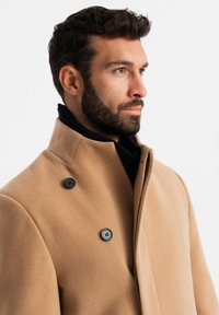 Helle Wolljacke mit erhöhtem Kragen, vorne mit zwei schwarzen Knöpfen. Der Stoff hat eine glatte Textur und einen sauberen, maßgeschneiderten Schnitt.
