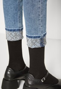 Jeans in denim blu chiaro con orli decorati, caratterizzati da un motivo a diamante e dettagli in argento. Abbinati a calzini neri e scarpe nere con tacco.
