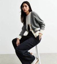 Grijze cardigan met crèmekleurige accenten en gouden knopen, gecombineerd met zwart gestreepte wijde broek en witte sneakers. Soepele texturen, casual uitstraling.