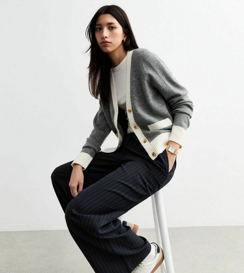 Cardigan grigio con dettagli crema e bottoni dorati, abbinato a pantaloni a gamba larga a righe verticali neri e sneakers bianche. Texture morbide, look casual.