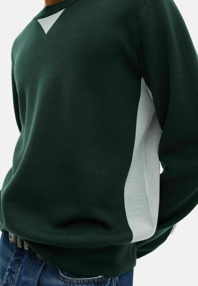 Maglione verde scuro con pannelli laterali bianchi e un piccolo logo ricamato, indossato con jeans blu.