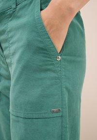 Zelené bavlněné kalhoty se bočním kapsou, malým kovovým logem a viditelnými švy. Měkká textura a uvolněný střih.