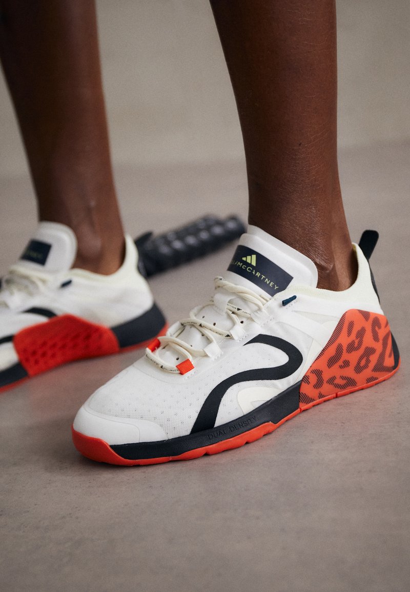 White athletic shoes with a black and orange leopard print accent. Features a textured fabric upper, rubber sole, and lacing system.