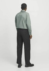 Cămașă verde cu mâneci lungi, guler și manșete cu nasturi, purtată cu pantaloni negri. Țesătură netedă, cu o croială lejeră și detalii minime.