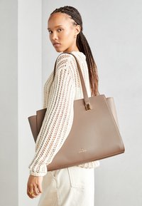 Tote bag in light brown leather, structured shape, with a gold accent. Attached shoulder straps and smooth texture, carried on the shoulder.
