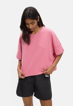 Kobieta nosząca luźny różowy pasiasty t-shirt i czarne szorty, trzymająca brzeg koszulki obiema rękami i patrząca w dół.