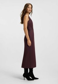 Lang, ærmeløs kjole i dyb burgunderrød stof med en tætsiddende overdel, flydende skørt og subtile sidesømme detaljer; kombineret med sorte stiletstøvler.