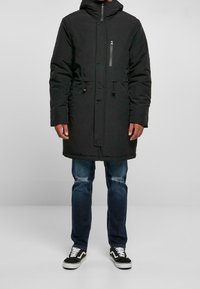 Černá péřová parka s kapsou na zip na hrudi, stahovacími šňůrkami v pase, kapucí, dlouhými rukávy a rovný střih, doplněná o rumměné modré džíny a tenisky.