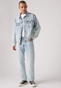 Giacca di jeans azzurro chiaro con due tasche sul petto, abbinata a jeans azzurro chiaro e sneakers nere basse. Texture liscia, design casual.