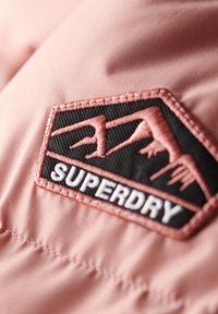 Un tessuto rosa presenta una toppa nera texturizzata con cuciture bianche. La toppa ha un design di montagna contornato e la parola "SUPERDRY."