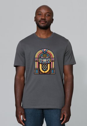 Mann trägt graues T-Shirt mit farbigem Vintage-Jukebox-Motiv, steht vor schlichtem hellgrauem Hintergrund.