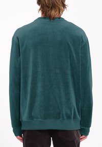 Un sudadera verde de tela suave con cuello redondo, puños acanalados y dobladillo. Presenta una textura lisa y un corte relajado, sin logotipos visibles.