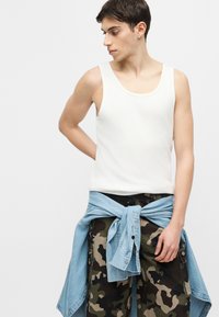 Giovane uomo che indossa una canotta bianca, pantaloni cargo mimetici e una camicia di jeans azzurro chiaro annodata in vita, che guarda verso sinistra.