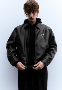 Zwarte leren bomberjack met ritssluiting, voorzien van een zilveren embleem op de borst. Gedragen over een zwarte hoodie met een gestructureerde afwerking.