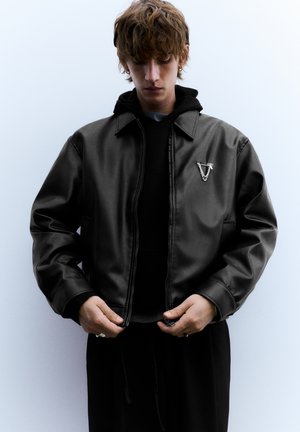 Crna bomber jakna od kože s patentnim zatvaračem, sadrži srebrni grb na prsima. Nosi se preko crne dukserice s kapuljačom, s teksturiranim završetkom.