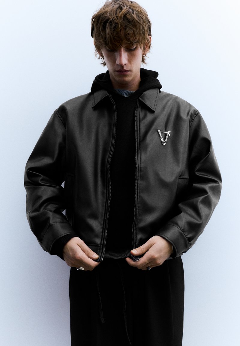 Zwarte leren bomberjack met ritssluiting, voorzien van een zilveren embleem op de borst. Gedragen over een zwarte hoodie met een gestructureerde afwerking.
