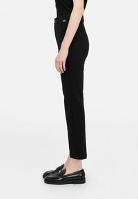 Pantaloni sartoriali neri con taglio dritto, caratterizzati da un piccolo dettaglio con cerniera dorata. Indossati con mocassini neri, vista laterale che mostra una silhouette sottile.