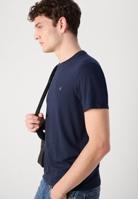 Tricou bleumarin cu mâneci scurte, decolteu rotund, textură moale și un logo mic pe piept. Persoana ține o curea de geantă neagră.