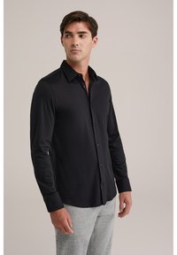 Juoda ilgomis rankovėmis marškiniai iš lygaus audinio, su sagomis priekyje, smailu apykakle ir susagstomais rankogaliais, dėvimi su šviesiai pilkomis kelnėmis.