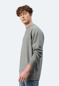 Felpa grigia con polsini a costine e scollo rotondo. Tessuto morbido, veste comodo e con dettagli laterali visibili. Indossata con jeans azzurri chiari.