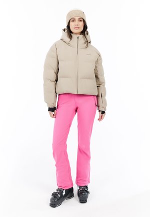 Femme portant une doudoune beige, un bonnet en tricot beige, un pantalon de neige rose vif et des bottes de ski noires, se tenant devant un fond blanc.
