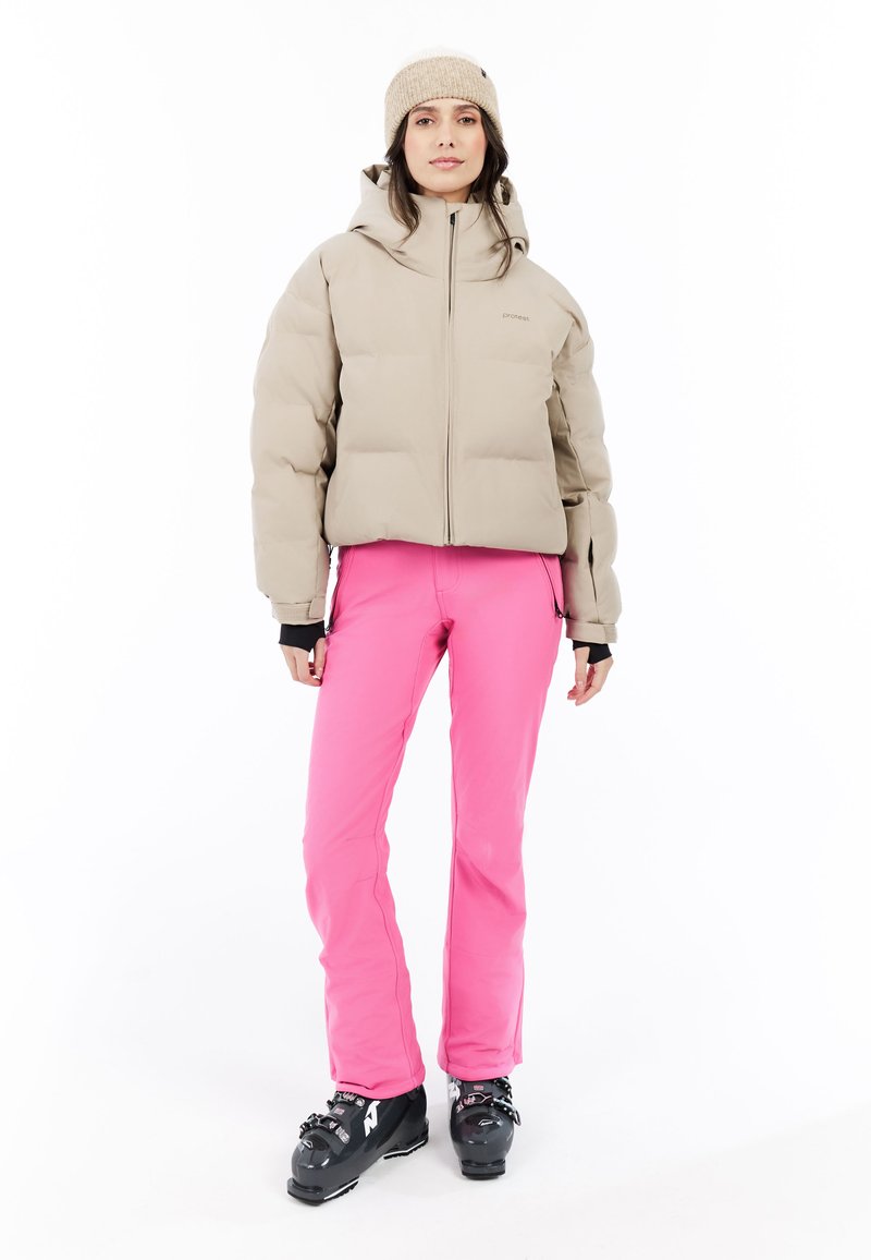 Vrouw in een beige pufferjack, beige gebreide muts, felroze skibroek en zwarte skiboots, staand tegen een witte achtergrond.