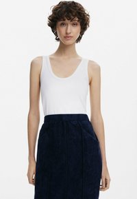 Ung kvinna med kort lockigt hår som bär en vit ärmlös topp och en mörkblå högmidjad kjol med fickor, står mot en enhetlig bakgrund.