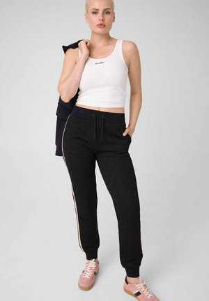 Femme portant un crop top blanc sans manches, un pantalon de jogging noir avec des bandes latérales, des baskets roses, tenant une veste noire sur l'épaule, debout devant un fond uni.