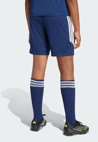 Σκούρες μπλε ποδοσφαιρικές βερμούδες με λευκές ρίγες, υφασμένο ύφασμα, σετ με σκουρόχρωμες μπλε καλτσες έως το γόνατο με λευκές ρίγες και μαύρα αθλητικά παπούτσια με τακούνια.