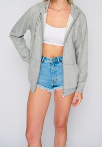 Felpa con zip di colore grigio chiaro, dal tessuto morbido e con cappuccio regolabile tramite cordino. Abbinata a pantaloni corti di jeans blu con orli sfilacciati.