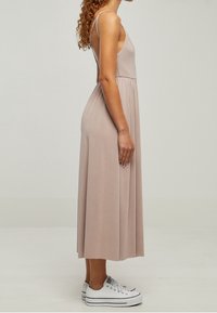 Femme portant une robe midi beige sans manches avec de fines bretelles et des baskets blanches à plateforme, debout de profil sur un fond uni.