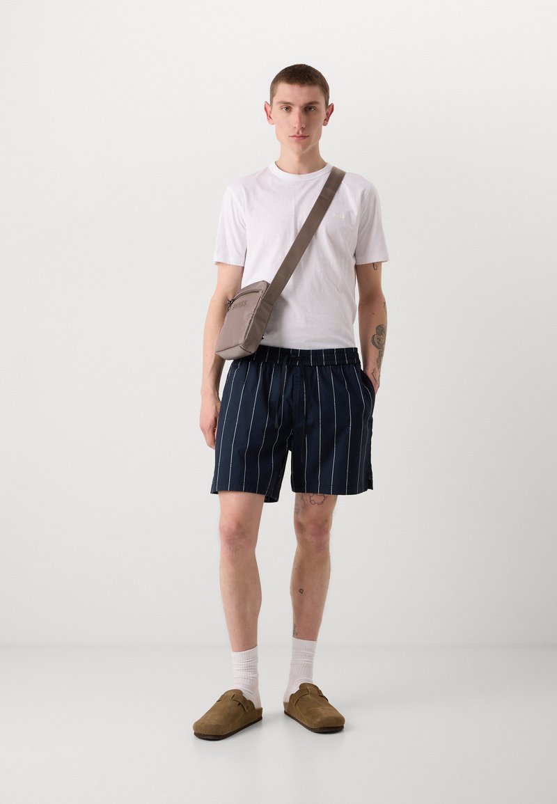 Weißes T-Shirt, marineblau gestreifte Shorts, beiger Umhängetasche. Modelliert mit braunen Slippern und weißen Knöchelsocken, vor einem schlichten Hintergrund stehend.