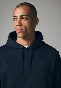 Sudadera con capucha de color azul marino, hecha de material suave, con cordones, agujeros para cordones en tono plateado y un pequeño logo bordado en el pecho.