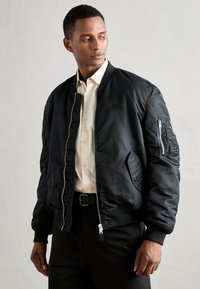Schwarze Bomberjacke aus glattem Stoff, mit einem Reißverschluss vorne, gerippten Bündchen und einer Brusttasche mit Reißverschluss.