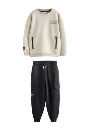 Beige langærmet sweatshirt med sorte lynlåslommer og sorte joggebukser med cargolommer og snøre i taljen.