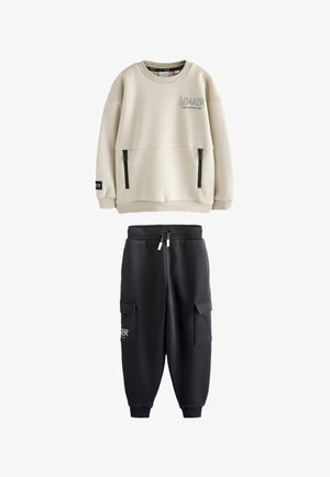 Beige langærmet sweatshirt med sorte lynlåslommer og sorte joggebukser med cargolommer og snøre i taljen.