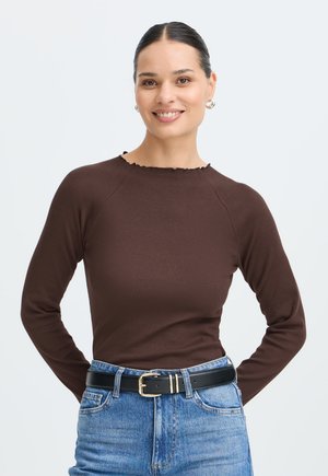 Femme souriante aux cheveux foncés attachés, portant un haut marron à manches longues, un jean bleu et une ceinture noire sur un fond clair uni.