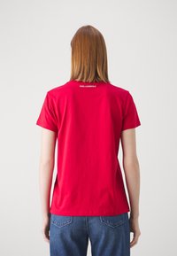 Persona con pelo hasta los hombros, vistiendo una camiseta roja de Karl Lagerfeld y jeans azules, de espaldas contra un fondo simple.