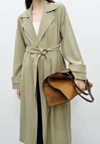 Długi, jasnooliwkowy płaszcz z przewiązanym w talii paskiem, z kołnierzem z klapami i szerokimi rękawami. W jednej ręce trzymana jest brązowa skórzana torba typu tote.