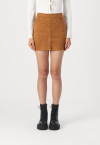 Mini jupe en daim marron avec fermeture à boutons sur le devant, deux poches latérales et une silhouette ajustée, assortie à des bottines noires à lacets.