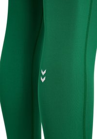 Zielone legginsy sportowe wykonane z gładkiego materiału, z białym logo w kształcie chevronu na udzie i kontrastowym ściegem wzdłuż szwów.