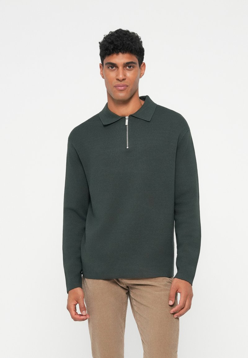 Mørkegrønn genser med glidelås i kragen og lange ermer, laget av teksturert strikket stoff, kombinert med beige cordfløyelsbukser.