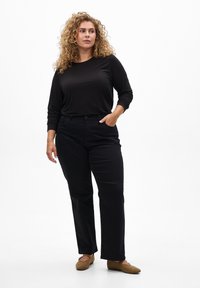 Svart långärmad topp i kombination med svarta byxor. Toppen är gjord av ett mjukt tyg, och byxorna har rak passform. Bruna skor med platt sula används.